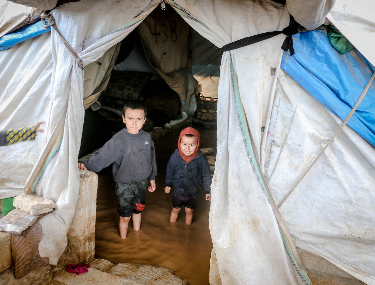 services-04 Two young children stand in a flooded tent in Idlib, Syria, highlighting the hardships in refugee camps.