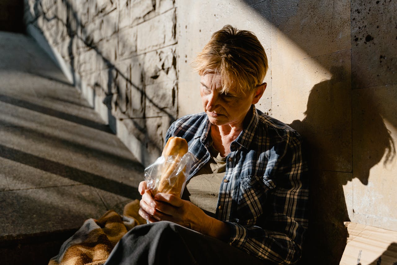 services-02 A senior woman holding and eating bread while seated against a sunlit wall, casting a thoughtful shadow.